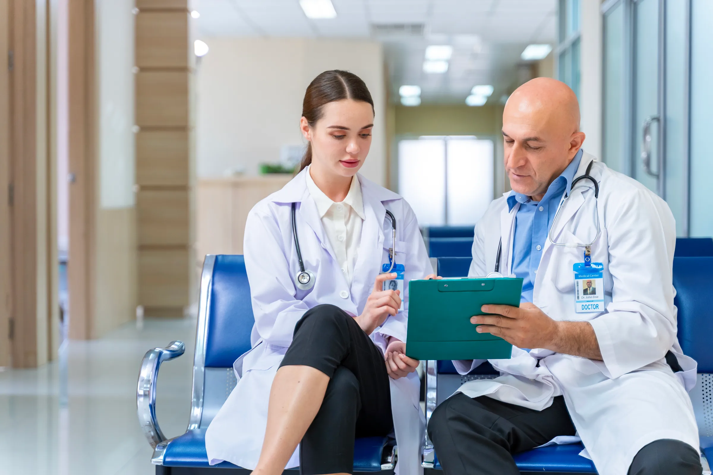 Two doctors wearing white coats and stethoscopes reviewing documents on a clipboard seated in a hospital.